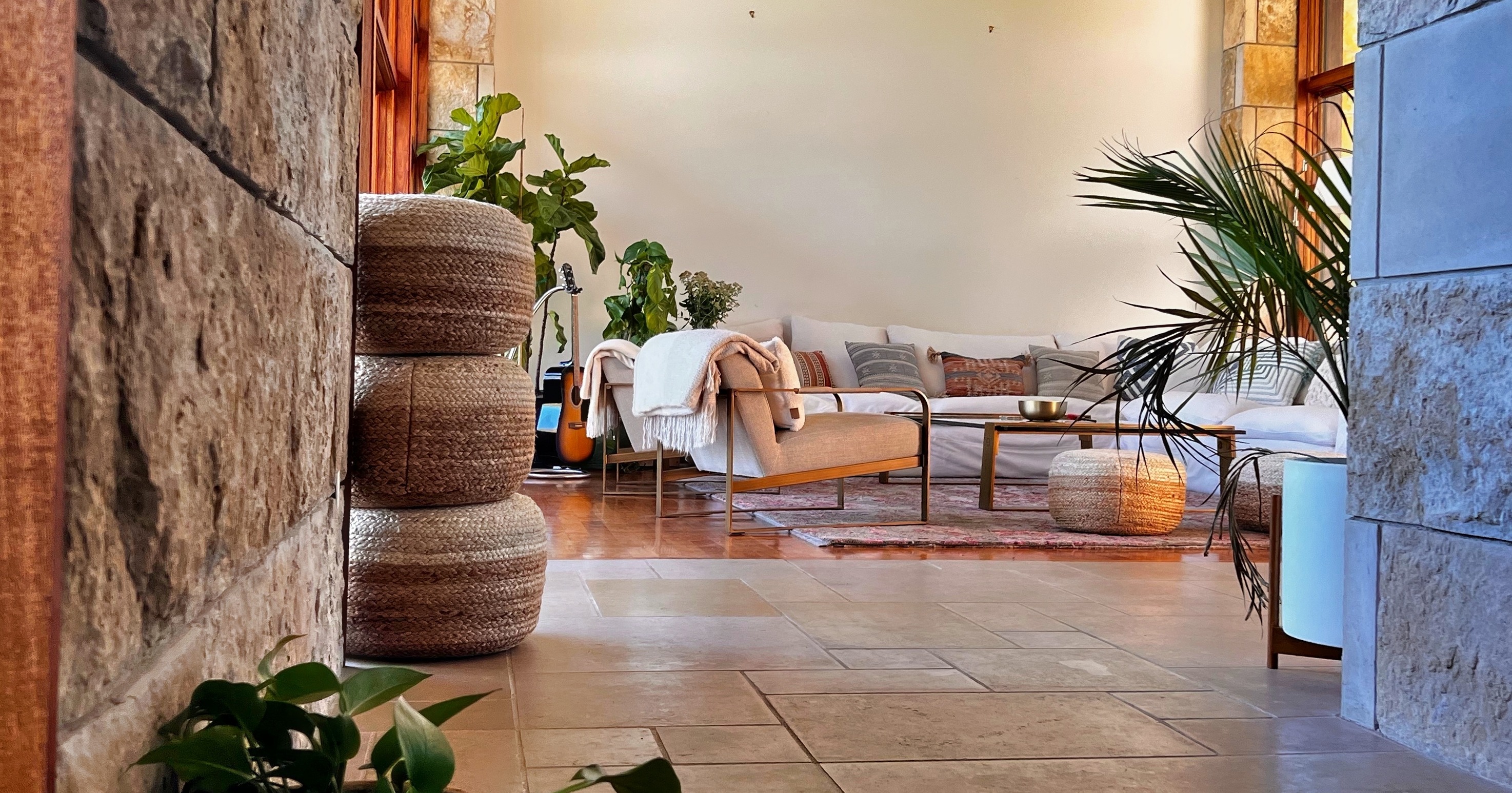 A sunlit indoor lounge with a guitar, woven poufs, and plants beside the stone-walled entrance to the main sitting room