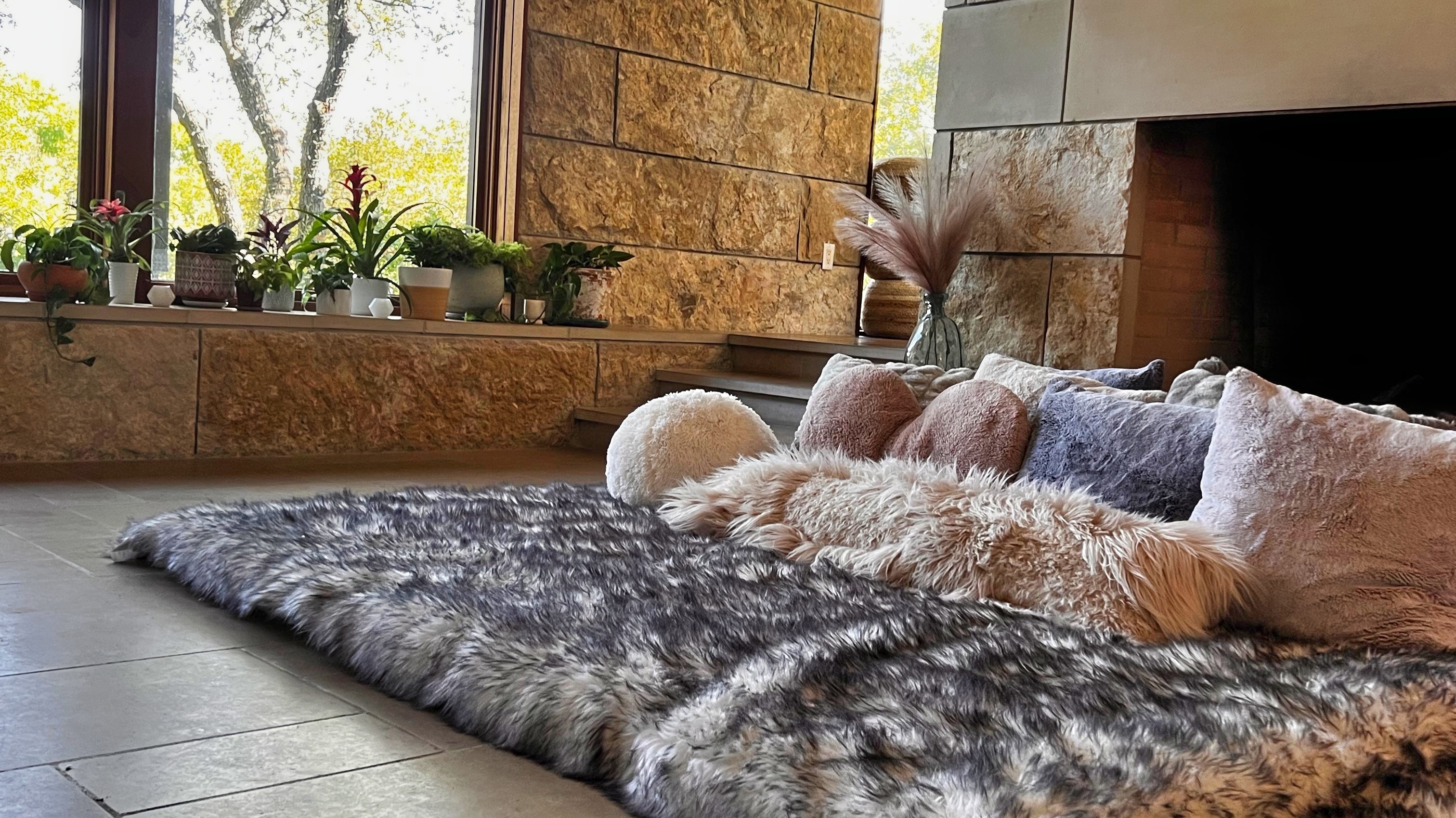 A plush faux-fur floor lounge with pillows in front of the stone fireplace, plants along the windowsill behind it